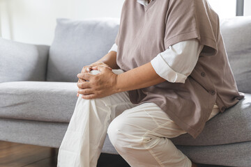 Body arthritis muscles stiff problem, aged woman suffering on leg, pain knee from disease chronic, hand massage rubbing joint injury, mature hurt or sore sitting on sofa at home. Health care people.