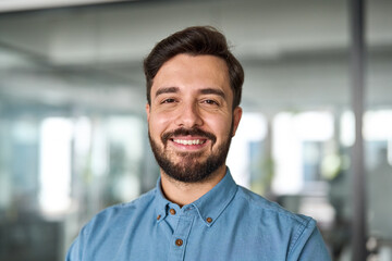 Obraz premium Confident happy young smiling Latin business man leader looking at camera standing in office. Successful happy businessman manager, professional executive or male entrepreneur, close up portrait.