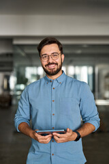 Happy young professional Latin business man looking at camera standing at work using tablet....