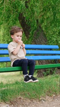 Small kid sits on the bench on a very windy day. Baby boy eating fruit from the doy pack. Vertical video.