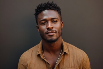 Obraz premium A young Black man with a short, textured hairstyle, wearing a brown shirt, gazes directly at the camera with a calm expression.