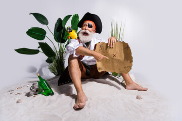 Senior pirate in buccaneer costume on sandy beach with treasure map and parrot, explores adventure with tropical plants and festive tone