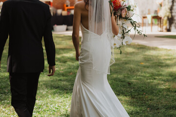Naklejka premium Elegant Bride and Groom Walking Hand in Hand During Outdoor Wedding Ceremony with a Flowing Bridal Gown and Stunning Floral Bouquet