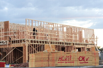 Two-story wood-frame house under construction with exposed studs, roof trusses, and stacked lumber marked for site use, as workers install framing components during residential build phase