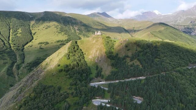 Aerial view of Gergeti Trinity Church in majestic Caucasus Mountains with serpentine road winding through dense spruce forest and leading to it, near Stepantsminda in Georgia.