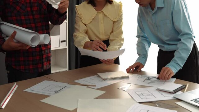 Architect and safety engineer discussion about building blueprint displaying on laptop on table with architectural document and wooden block scatter around at modern office. Closeup. Delineation.