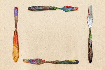 A frame made of four dirty, colorful palette knives arranged on a natural canvas background.