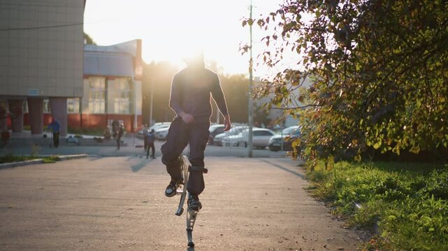 Front shot of man wearing gray hoodie and dark gray pants jumping on spring stilts on paved road in residential area surrounded by houses during sunset, with serious expression