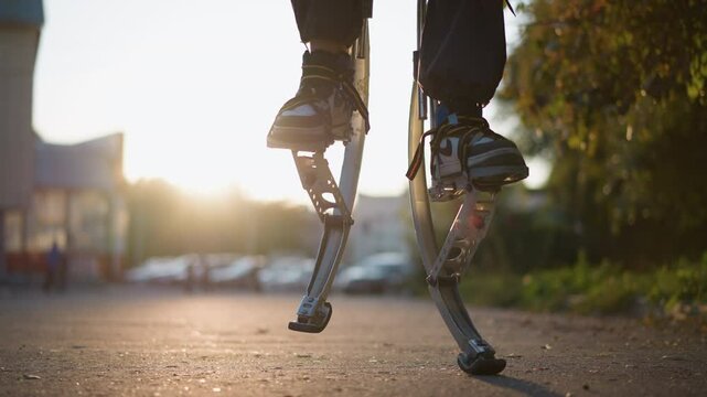 Close-up of a person's leg walking on spring stilts with dark pants and sneakers. Background shows a blurred suburban area with greenery, buildings, and vehicles under a warm sunset glow. Low-angle