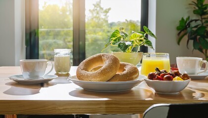 Indoor brunch table with sunlight shining through a glass window, buttered bagels and bowl of fresh fruit placed next to coffee cups