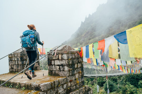 Young female trekker crossing river over Suspension Bridge decorated with multicolored Tibetan Prayer flags hinged over gorge near Lukla. Everest Base Camp route, Sagarmatha National Park, Nepal