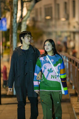 Obraz premium A young Chinese couple in their 20s walking along a riverside sidewalk in the Bund, Hongkou District, Shanghai, People's Republic of China, on December 1, 2024, during the cold season.