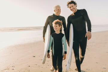 Three generations of surfers walking on the beach with surfboards at sunset