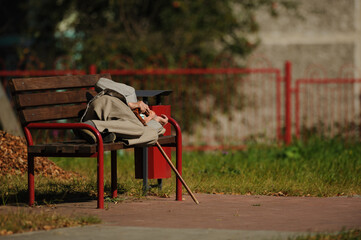 A elderly man in a gray suit, gray cap, gray shoes and with a cane lies on a bench in the street. 