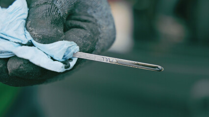 Checking Oil Level with Dipstick. A close-up view of a gloved hand holding an oil dipstick marked with the word FULL, while wiping it clean with a blue cloth, against a blurred background.