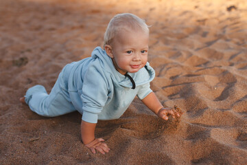 Fototapeta premium Happy toddler in blue hoodie crawls along beach in sand on sunny day
