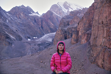 A young girl in bright clothes is sitting on a rock and smiling in the mountains against the background of rocks and snowy peaks. Trekking in the mountains in summer
