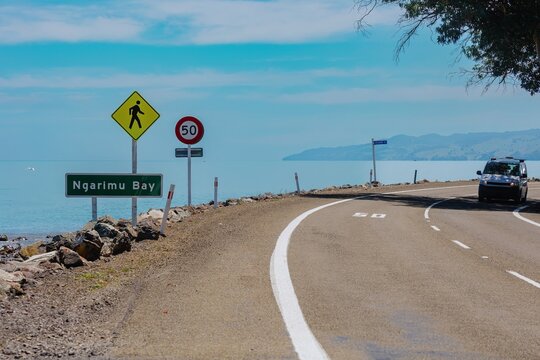 Coastal road with speed limit sign, pedestrian crossing, and a van driving along Ngariimu Bay. Safe travel. NGARIMU BAY, THAMES, COROMANDEL PENINSULA, NEW ZEALAND