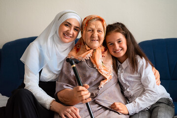 Portrait of muslim grandmother, mother and granddaughter hugging with love on the sofa smiling and looking at the camera. family of three female generations.