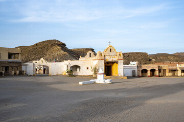 Charming Southwestern Village: Rustic Adobe Buildings, Small Church with Cross, and Quaint Central Courtyard Set Against Rocky Hills and Blue Sky, Capturing Traditional Mexican Architecture and Herita