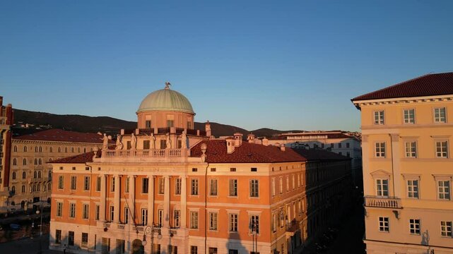 aerial view of downtown trieste (historic buildings on Mediterranean coast canal, famous buildings) boats in marina, cars driving road (sunset golden hour) red roof piazza palazzo footage drone italy