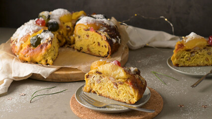 Festive fruitcake with powdered sugar, adorned with colorful candied fruits, served on a wooden...