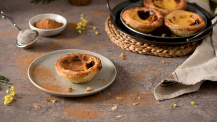 Pastel de Nata on a plate with cinnamon sprinkled around, surrounded by a sieve, a bowl of cinnamon, and more tarts on a woven mat