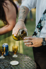 Close-Up of a Stylish Guest Pouring Pickle Juice into a Vintage Glass at an Outdoor Garden Party with Rustic Decor and Unique Drink Presentation