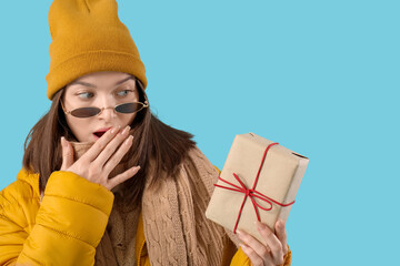 Shocked young woman in winter clothes with Christmas gift on blue background, closeup