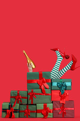 Woman in Christmas stockings holding champagne behind stack of presents on red background