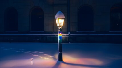 Snow-covered lamppost illuminated by rainbow light near dark building