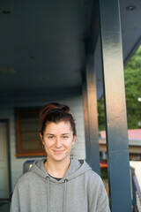 A casual portrait of a young woman wearing a grey hoodie, standing on a porch with a relaxed expression. The background features a mix of indoor and outdoor elements, including greenery and buildings