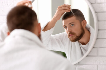 Fototapeta premium Young man using serum for hair growth near mirror in bathroom. Hair loss concept