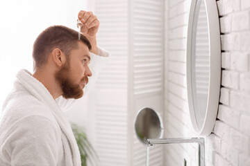 Young man using serum for hair growth near mirror in bathroom. Hair loss concept