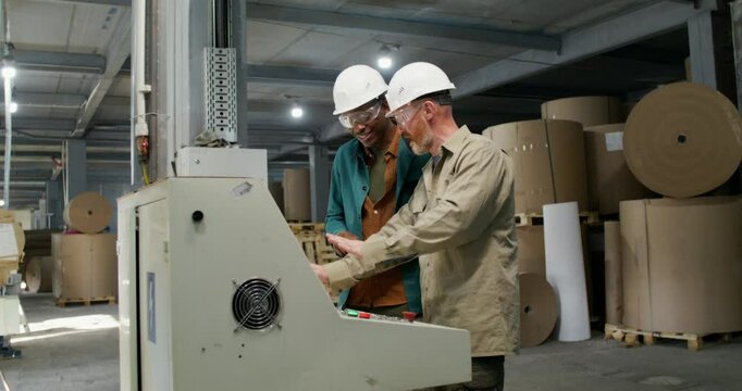 Two men in protective helmets and glasses are talking standing near the machine control panel at a paper mill