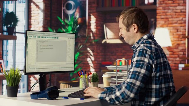 Man in home office stretches back while looking at UI on PC screen, feeling fatigued while executing programming code. IT developer feeling backpain, relieving pain, writing and testing code, camera B