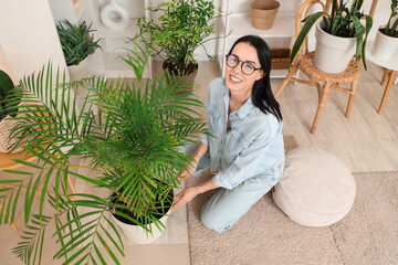 Beautiful young woman with gardening rake taking care of houseplant at home