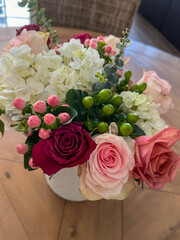 A stunning floral arrangement featuring white hydrangeas, deep red and blush pink roses, green and pink berries, and eucalyptus stems in a soft, natural light setting on a wooden table.