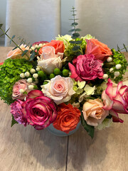 A striking floral centerpiece featuring pink, orange, and blush roses, green hydrangeas, white berries, and eucalyptus sprigs, arranged elegantly in a vase on a light wooden table.