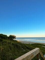 Obraz premium A wooden railing frames a serene view of the beach, bordered by grassy dunes and a calm ocean under a vast, cloudless blue sky. A peaceful coastal landscape at its finest.