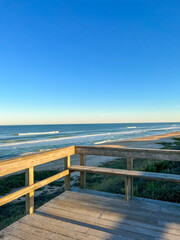 Obraz premium A wooden beach lookout overlooks serene ocean waves, a golden sandy shore, and lush dunes under a clear blue sky, capturing a peaceful coastal escape.