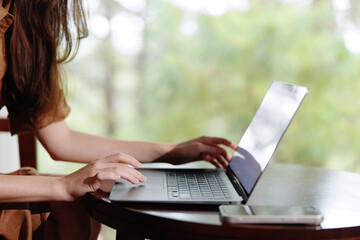 Fototapeta premium A woman working on a laptop at a wooden table, showcasing focus and productivity in a tranquil outdoor setting with blurred greenery in the background