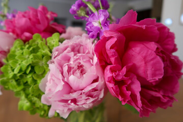 A bright floral arrangement featuring a striking magenta peony, soft pink blooms, green hydrangeas, and delicate purple flowers, beautifully arranged with a softly blurred background for depth.