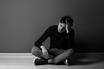 Black and white portrait of depressed young man sitting near dark wall