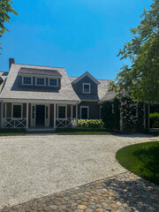 A sunlit, elegant home with a gravel driveway, cobblestone accents, lush greenery, and blooming...