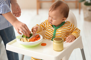 Mother feeding her little baby with vegetables in high chair at home