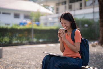 Happy young asian student enjoying a coffee break while studying on her laptop, sitting on a bench outdoors under the warm sun, embracing a cheerful moment of relaxation