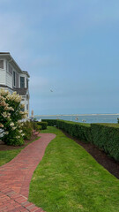 A picturesque red brick pathway lined with vibrant hedges and flowering shrubs leads to a coastal view of boats gently anchored in calm waters under a soft blue sky.