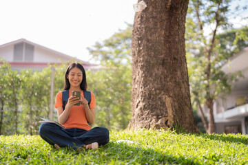 Happy asian female student sitting with crossed legs under tree on university campus lawn using...