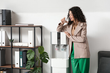 Beautiful businesswoman drinking water near cooler in office